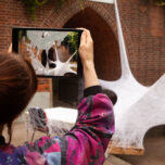 A person holds a tablet in front of a sculpture in the garden. The screen shows the same sculpture, but adding some digitially rendered sculpturs to the image