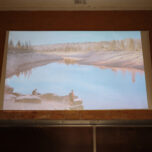 Foto of the projection screen. On the screen an image of a lake, and a person standing next to it, gazing over the water
