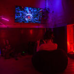 In one of the cages in Bärenzwinger. A visitor sits on a beanbag and plays a video game. The room is immersed in red and blue light. House plants decorate the scene. The screen shows a still of the game, a blurry futurist environment suggesting that the player’s avatar moves at a great speed.