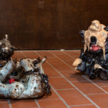 3 glasierte Ton-Skulpturen von Hunden die Metallketten um ihren Körper tragen. Die 2 Hunde auf der linken Seite des Bildes scheinen auf dem Boden zu rollen, der Hund rechts sitzt aufrecht, lächelt und hat einen großen Haufen Scheiße auf dem Kopf.