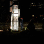 Noch ein Foto der Installation im Garten. Die Skulptur, bei Nacht fotografiert, wird von innen erleuchtet. Ein:e Besucher:in steht davor und betrachtet sie aufmerksam.