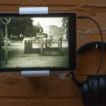 Foto eines iPads, auf dem ein Schwarzweißfilm läuft. Die Kamera fokussiert auf den Eingang einer öffentlichen Toilette. Auf einem Schild über dem Eingang steht: Männer. Autos fahren auf einer Straße neben dem Schild vorbei.